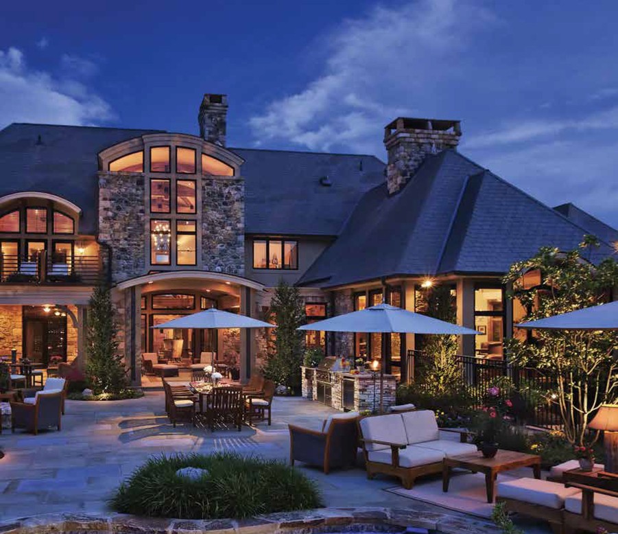 A large stone house with landscape lighting and an outdoor patio and pool space.  